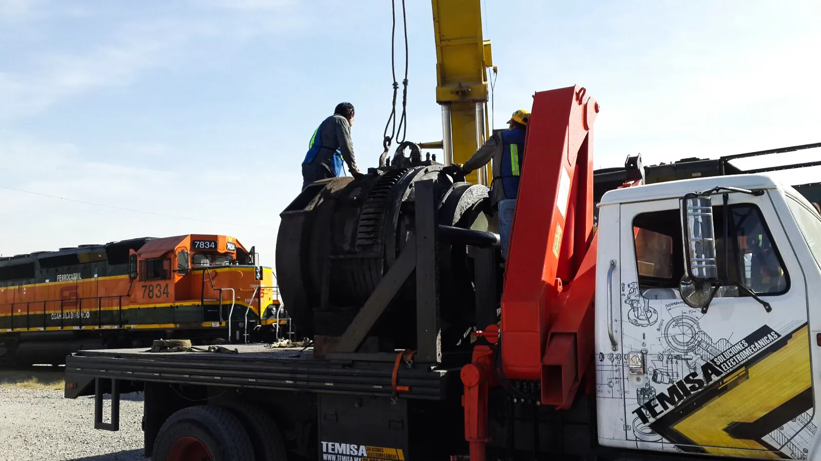 Motor de tracción cargado en la grúa de TEMISA junto a una locomotora diésel-eléctrica en el patio ferroviario