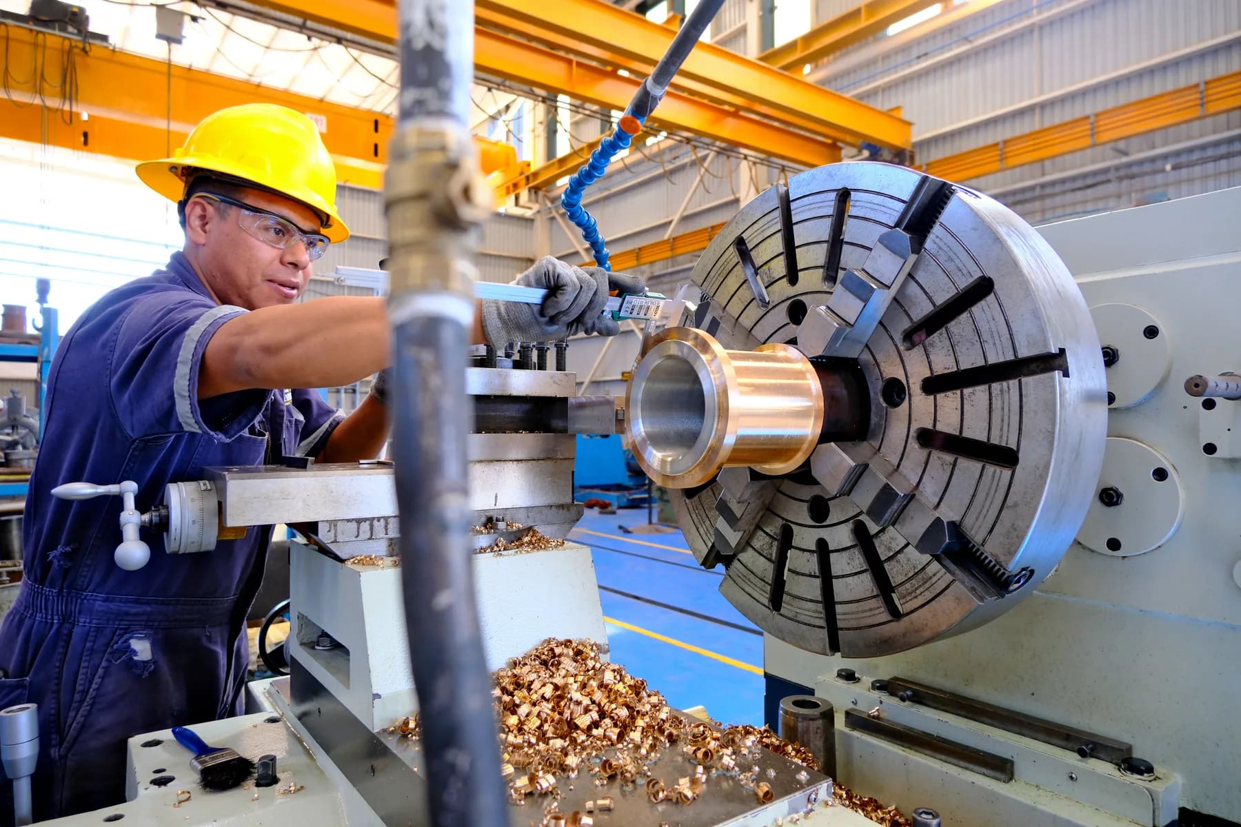 Técnico maquinando y midiendo con calibrador un buje de bronce sujeto en el plato del torno, con viruta de bronce sobre la bancada